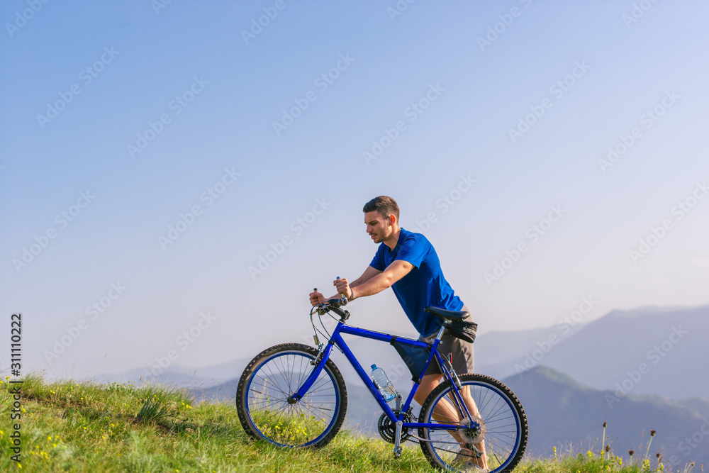 Tired fit mountain biker pushing his bike uphill at the top of the mountain on a sunny day with amazing view on a blue river.