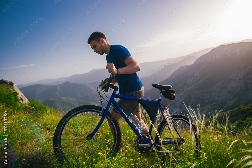 Perspective of a fit mountain biker pushing his bike uphill with amazing view on a forest, river and mountains in the background. Amazing green nature at sunset.