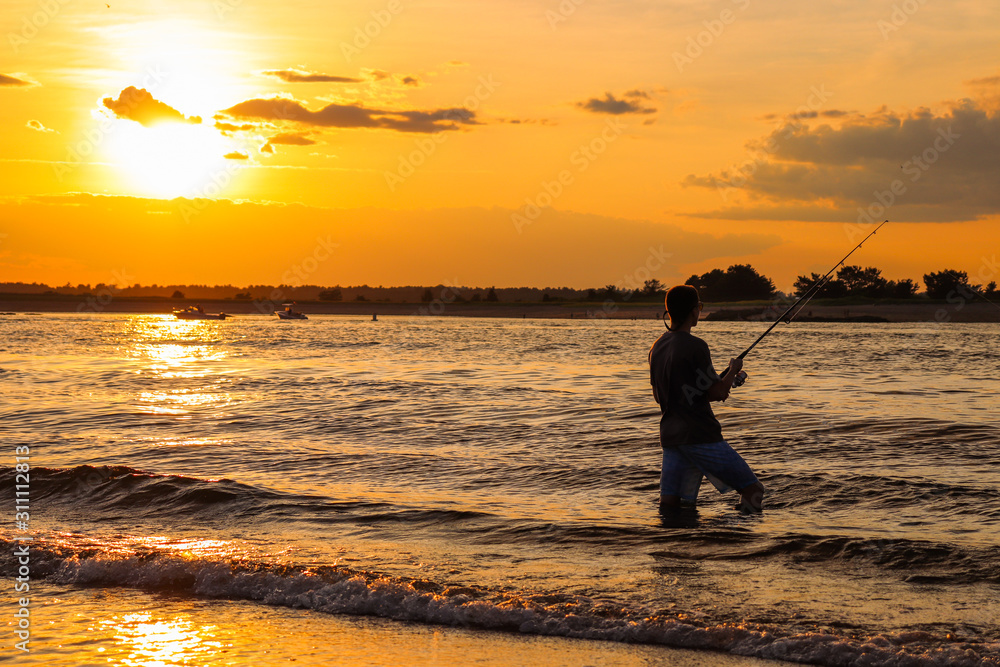A young man that is fishing during sunset on a beach