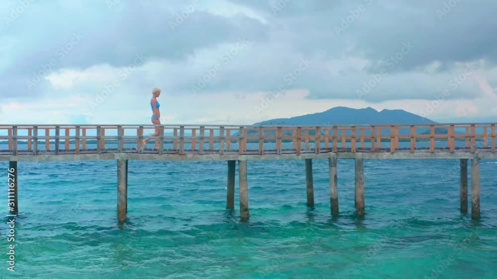 Woman walks on wooden pier in a summer day. Tourist sexy girl in blue bikini enjoying European summer holiday travel vacation. Asia adventure. Cheerful girl with short blonde hair on a wooden pier. Woman walks on wooden pier in a summer day. Tourist sexy girl in blue bikini enjoying European summer holiday travel vacation. Asia adventure. Cheerful girl with short blonde hair on a wooden pier.