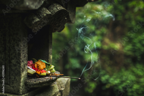 Traditional balinese Canang Sari offerings to gods and spirits with flowers, food and smoky aromatic sticks on dark green background. Indonesian culture and religion. Bali authentic travel concept. photo