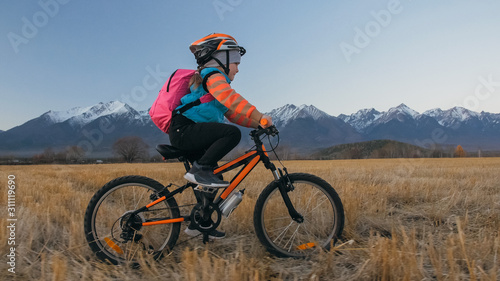 One caucasian children rides bike in wheat field. Little girl riding black orange cycle on background of beautiful snowy mountains. Biker motion ride with backpack and helmet. Mountain bike hardtail.