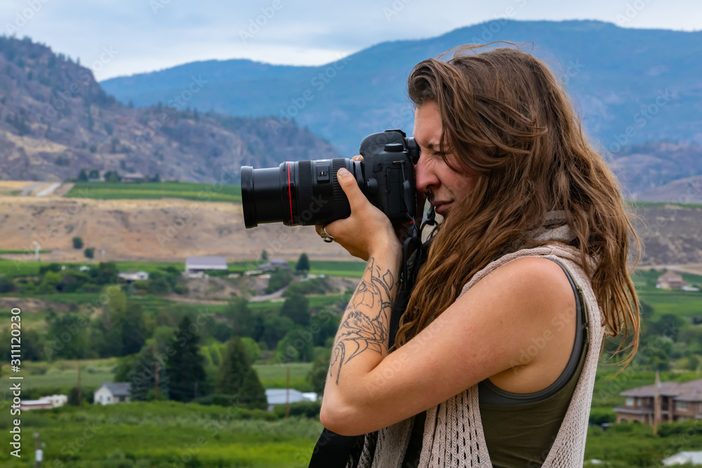 Young caucasian woman photographer with long hair and tattoo, on the hill taking pictures of vineyards farms, with her DSLR camera, close up side view