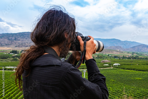 Young caucasian man photographer with long hair rear side view on the hill, taking pictures with a tele lens of vineyards farms with his DSLR camera