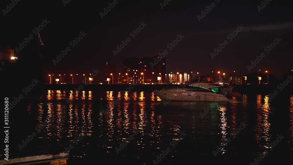 Luxurious modern private yacht and boat at the pier at night. Ship crossing skyline in the harbor in summer awesome nightly. Colorful lights of city glow shine reflected in water wave at nighttime.