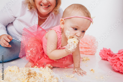 Birthday celebration: little girl eating cake with her hands on white background. child is covered in food. on floor among decoration: numbers 1, artificial flowers and white balls. Copy space photo