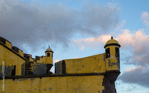Detail of the bright yellow brick and stone walls of Fort of Sao Tiago, located in the historical centre of Funchal, Madeira. Photo is taken during the sunset that colored the sky and clouds.
