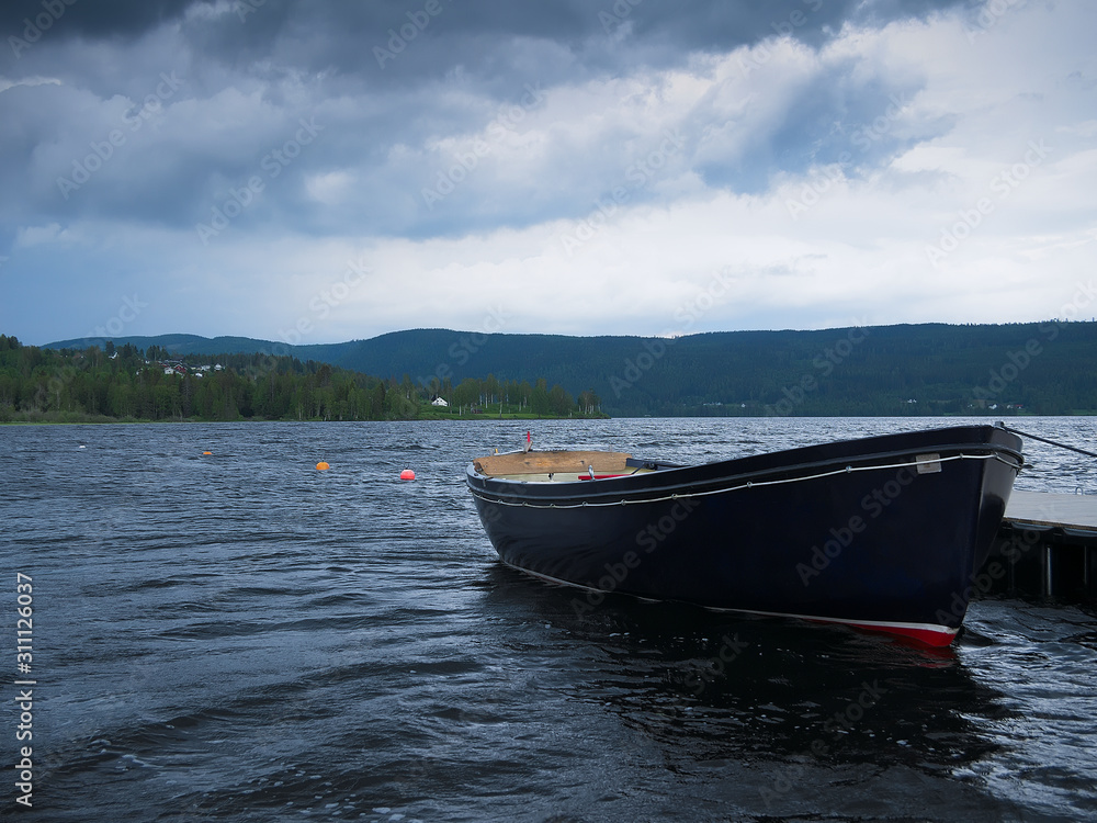 Dark row boat tied to a dock on beautiful lake Hurdalssjoen in Norway, with storm clouds overhead. Beautiful Europe Travel Scene.