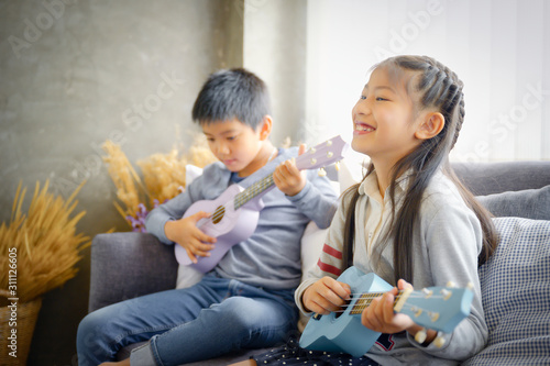 Happy elementary age Asian little kid is smiling  while playing a ukulele during a private music learning lesson at home photo