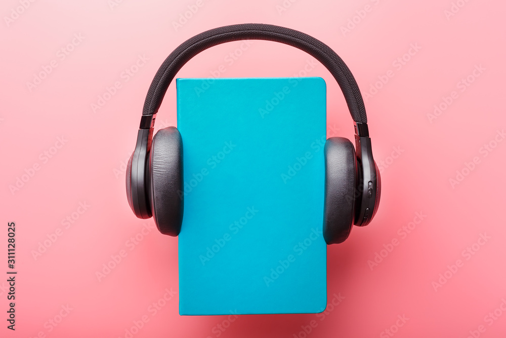 Headphones are worn on a book in a blue hardcover on a pink background, top view. Headphones are worn on a book in a blue hardcover on a pink background, top view.