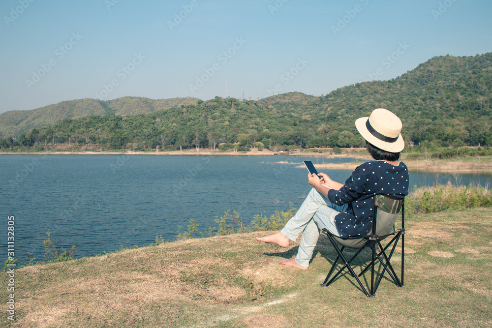 Vacation and Travel Concept : Asian woman wear hat and sitting relax on portable chair and playing smartphone nearly lake at National Park.