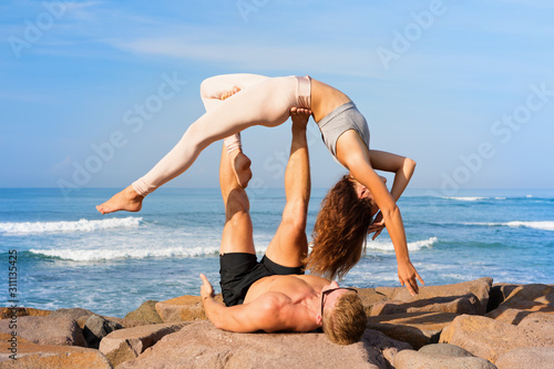 Fit young couple doing acro yoga at spa retreat on sea beach. Active woman balancing on partner feet, stretching at acroyoga pose. Healthy lifestyle. People outdoor sport activity on family vacation. photo