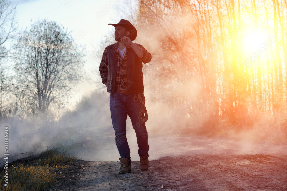 Fototapeta premium A man is wearing a cowboy hat and a loso in the field. American farmer in a field wearing a jeans hat and with a lasso in the smoke of a fire. A man walks through a burning field