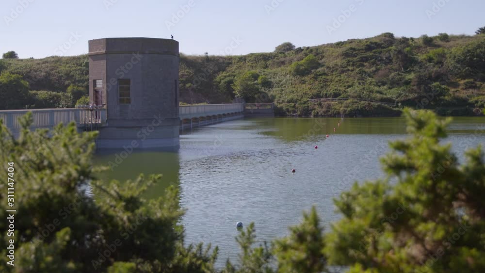 A steady long shot of a calm reservoir located between lush riverbanks that are connected by a concrete dam with a watchtower in the middle. A steady long shot of a calm reservoir located between lush riverbanks that are connected by a concrete dam with a watchtower in the middle.