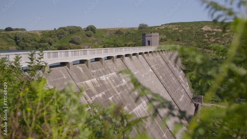 A steady wide shot of a concrete dam of a reservoir with a watchtower in the middle and located between two lush terrains. A steady wide shot of a concrete dam of a reservoir with a watchtower in the middle and located between two lush terrains.