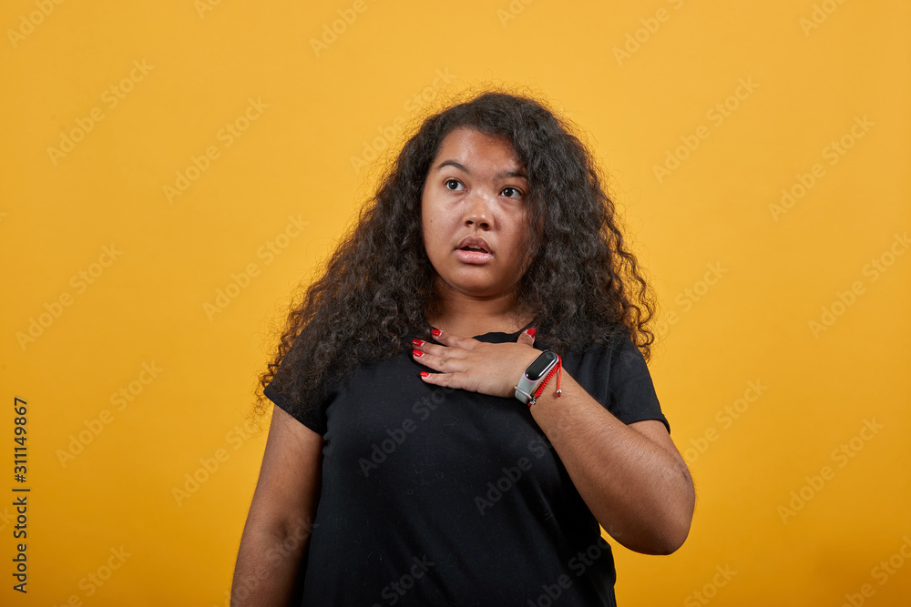 Shocked afro-american young woman with overweight keeping hand on chest, looking at camera over isolated orange background wearing fashion black shirt. People lifestyle concept. Shocked afro-american young woman with overweight keeping hand on chest, looking at camera over isolated orange background wearing fashion black shirt. People lifestyle concept.