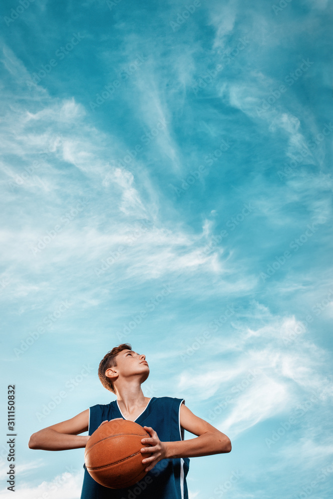 Fototapeta premium Sports and basketball. A young teen in a black tracksuit playing basketball on the school Playground. Cloudy blue sky on the background. View from the bottom. Vertical. Copy space