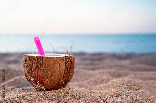 Coconut cocktail at the beach. Pink straw in coconut set on the sand against the blue sky and sea. Copy space, outdoors.