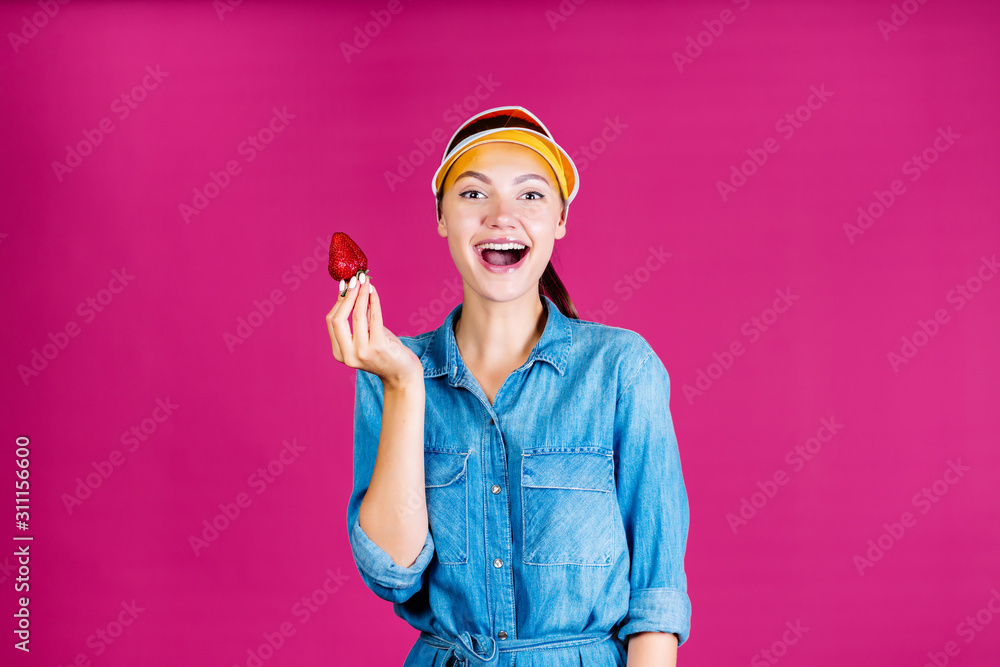 the seller the girl in the visor joyfully opened her mouth in an exclamation and holds the strawberry in her hand. fuchsia background the seller the girl in the visor joyfully opened her mouth in an exclamation and holds the strawberry in her hand. fuchsia background