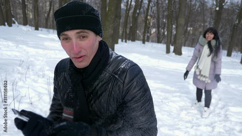 Handsome Young Man Talking On Smartphone. Girl Throws Snowball In Man's Back. Slow Motion Winter Sunny Day Handsome Young Man Talking On Smartphone. Girl Throws Snowball In Man's Back. Slow Motion Winter Sunny Day