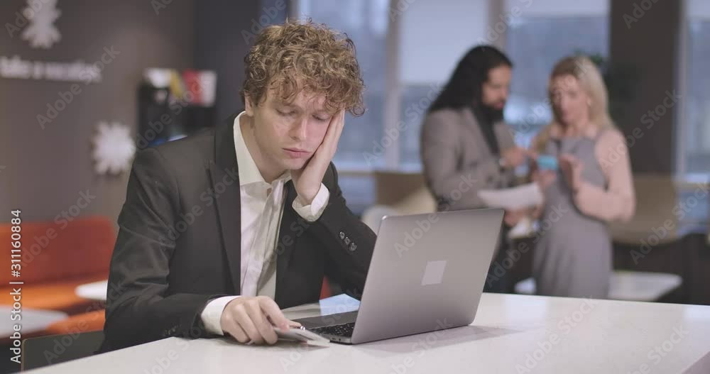 Portrait of tired Caucasian young man typing on laptop keyboard, looking at smartphone, and rubbing head. Exhausted male employee overworking in open space office. Cinema 4k ProRes HQ. Portrait of tired Caucasian young man typing on laptop keyboard, looking at smartphone, and rubbing head. Exhausted male employee overworking in open space office. Cinema 4k ProRes HQ.