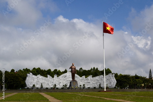 Pleiku, Vietnam - September, 2015: Wide square with bronze monument of communist leader Ho Chi Minh and high rise flagpole with national red flag with yellow star on background of blue cloudy sky