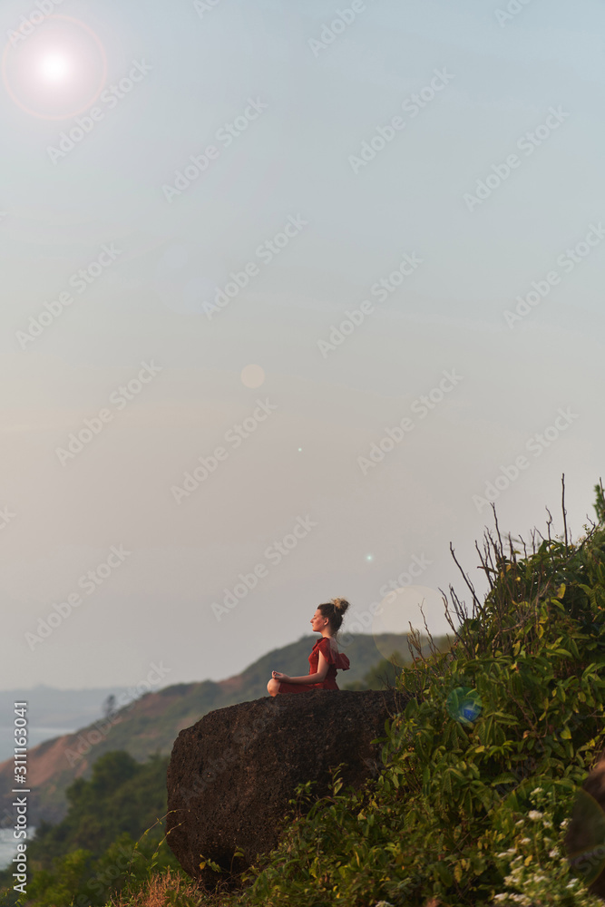 Fototapeta premium Young girl practices yoga in a red dress on a mountain in India