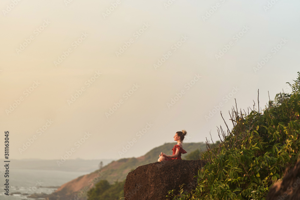 Fototapeta premium Young girl practices yoga in a red dress on a mountain in India
