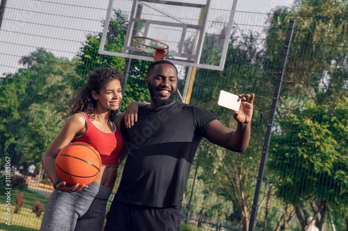 Outdoors Activity. African couple standing on basketball court boyfriend taking selfie on smartphone with girlfriend smiling happy