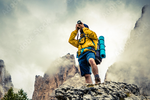 Young man hiker taking pictures on stone mountain with cloudy sky and fog. Yellow jacket, backpack, black beard and beanie. Traveling Dolomites, Italy.