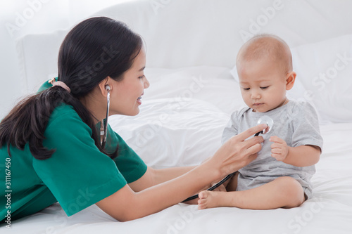 Asian toddler girl get sick examine by pediatrician doctor woman green uniform hold stethoscope  mother or nurse monitoring heart pulse rate adorable infant on bed in clinic  baby health care concept