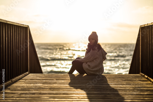 Pretty Young girl sitting on a bridge that goes to the beach with beautiful sunset on the sea