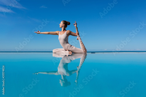 Charming woman yogi in overalls makes One-Legged King Pigeon, Eka Pada Rajakapotasana Pose by pool while relaxing at sea in sunny warm country. The concept of relaxation and better health. Copyspace