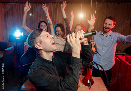 clubber man emotionally singing in microphone, wearing black shirt, have fun in karaoke bar, friends support him andjoining to him in the background