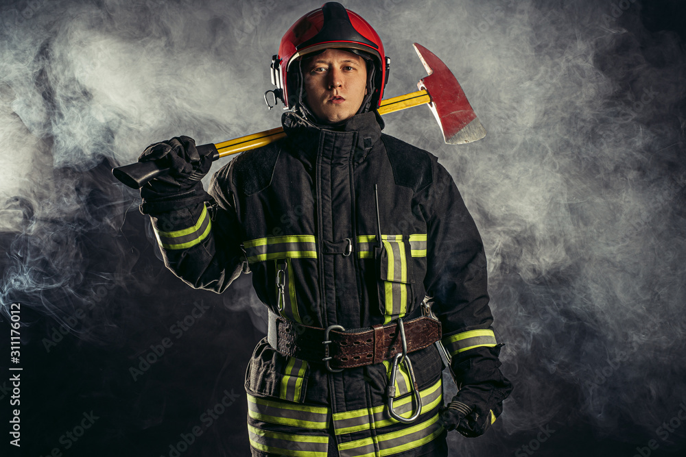 Fototapeta premium young caucasian handsome and strong fireman in uniform wearing helmet on head hold hammer in hands, going to save people from fire