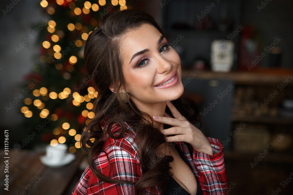 Beauty portrait of a beautiful sexy brunette girl in red checkered pajamas against the background of the New Year tree.