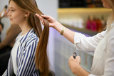 careful caucasian hairdresser holding client's hair in hands, going to cut off tips of hair, making hairdo to young girl client sitting on chair opposite of mirror in beauty salon