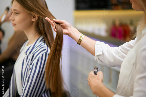 careful caucasian hairdresser holding client's hair in hands, going to cut off tips of hair, making hairdo to young girl client sitting on chair opposite of mirror in beauty salon