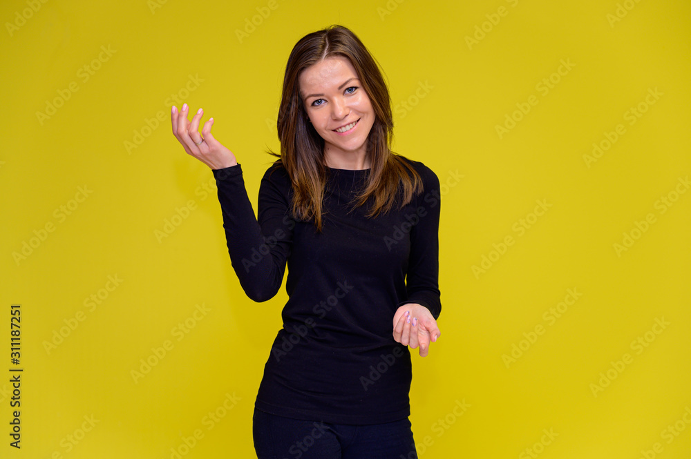 The concept of a smiling cute girl with positive emotions. Portrait of a young woman in a black T-shirt stands in different poses with long hair on a yellow background. Shows hands, talking. The concept of a smiling cute girl with positive emotions. Portrait of a young woman in a black T-shirt stands in different poses with long hair on a yellow background. Shows hands, talking.