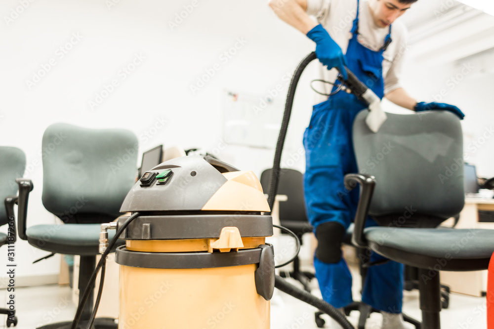 Young man in workwear and rubber gloves cleans the office chair with professional equipment. Young man in workwear and rubber gloves cleans the office chair with professional equipment.