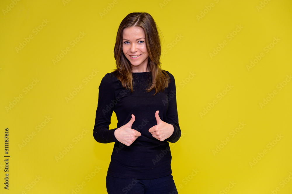 The concept of a smiling cute girl with positive emotions. Portrait of a young woman in a black T-shirt stands in different poses with long hair on a yellow background. Shows hands, talking. The concept of a smiling cute girl with positive emotions. Portrait of a young woman in a black T-shirt stands in different poses with long hair on a yellow background. Shows hands, talking.