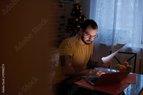 young hardworking man of caucasian appearance, with beard work on laptop at home, holding documents in hands