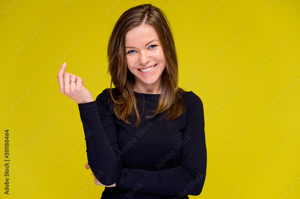 The concept of a smiling cute girl with positive emotions. Portrait of a young woman in a black T-shirt stands in different poses with long hair on a yellow background. Shows hands, talking. The concept of a smiling cute girl with positive emotions. Portrait of a young woman in a black T-shirt stands in different poses with long hair on a yellow background. Shows hands, talking.