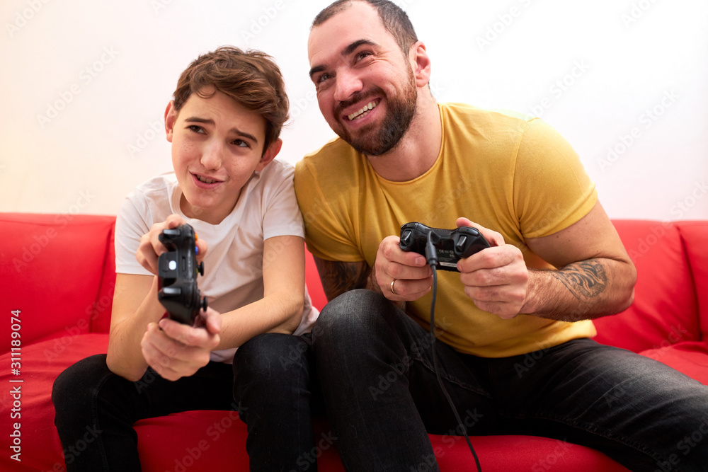Fototapeta premium young caucasian man with beard sit with son on sofa and play video game together, holding joysticks, wearing casual t-shirt. indoors