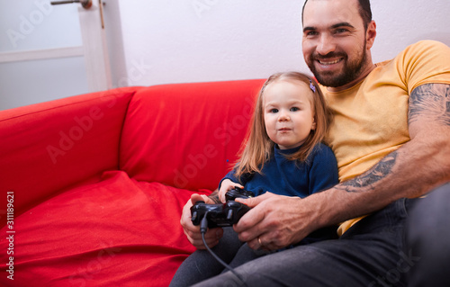 sweet adorable little daughter sit with dad in playing center, play video games sitting on reed sofa