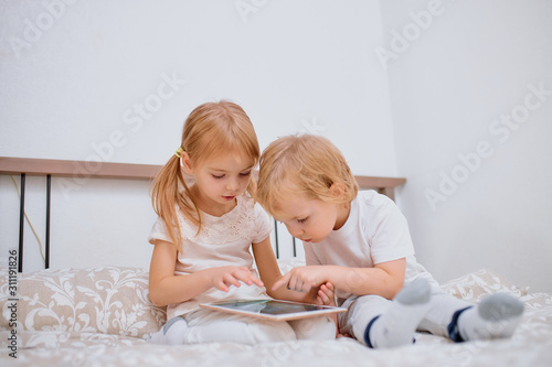 little caucasian children, boy and girl look at screen of laptop, playing or watching videos on modern gadget, wearing white casual clothes, sit on bed at home