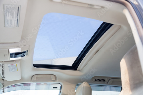 View of the ceiling of the car with a transparent glass hatch for airing, opening so that you can see the sky and clouds in the gray cabin of the vehicle. Sunroof on beige roof.