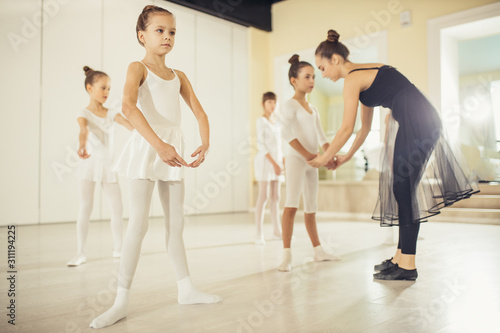 young caucasian ballerina in black suit teaching little children in white suits, practicing and coaching them in studio, correct body position