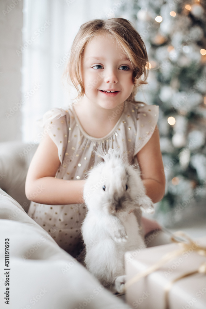Stock photo portrait of cute little girl in festive dress with lovely bunny in her arms sitting on couch. Unfocused decorated Christmas tree in background. Stock photo portrait of cute little girl in festive dress with lovely bunny in her arms sitting on couch. Unfocused decorated Christmas tree in background.
