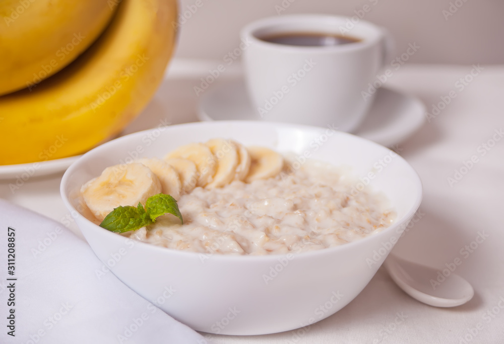 Oatmeal porridge with banana in the white bowl, cup of coffee on the white table. Health breakfast. Oatmeal porridge with banana in the white bowl, cup of coffee on the white table. Health breakfast.